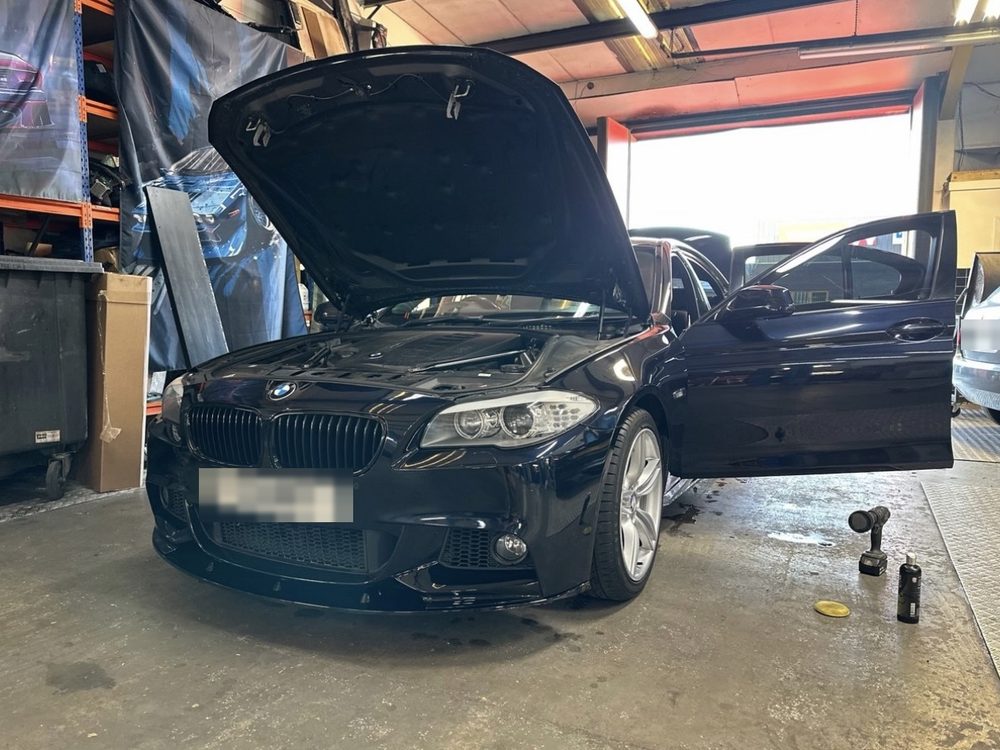 A dark blue BMW 5 Series with the bonnet up inside the workshop