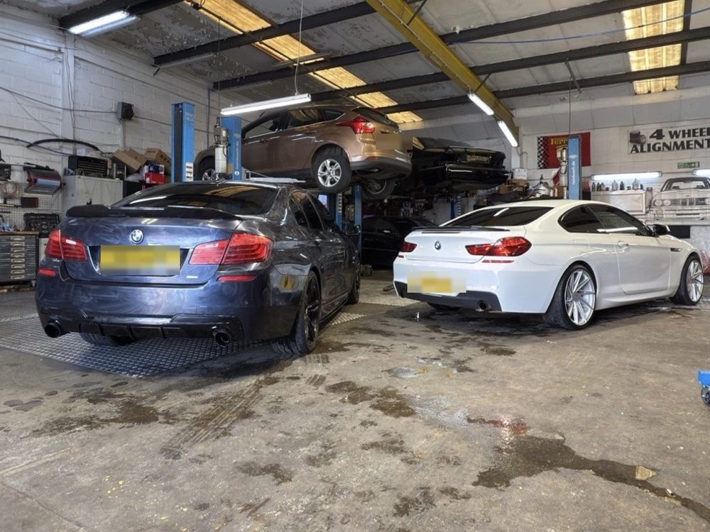Two BMWs on the workshop floor with a car up on the ramp behind them